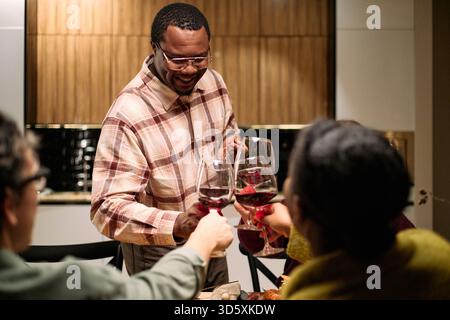 Homme noir d'âge moyen souriant tout en servant du vin et des verres de clinking avec divers groupes d'amis pendant le dîner d'action de grâce, mains visibles et visages de jeunes et de personnes d'âge moyen participant Banque D'Images