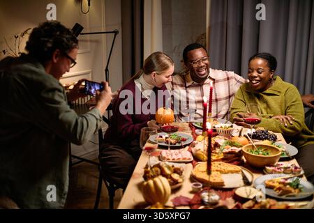 Groupe de jeunes adultes divers et amis d'âge moyen assis autour de la table partageant le dîner de Thanksgiving, souriant et riant tandis que l'homme photographiant le moment avec smartphone Banque D'Images