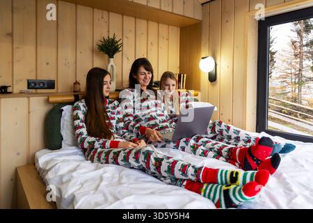 Famille en pyjama de Noël regardant ordinateur portable dans la cabine. Mère et filles portant un pyjama festif se relaxant dans une cabine en bois confortable, profitant du temps Banque D'Images