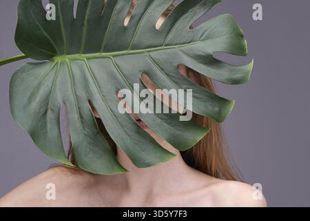 Mystérieuse jeune femme caucasienne partiellement cachée derrière une grande feuille de monstera sur fond gris Banque D'Images