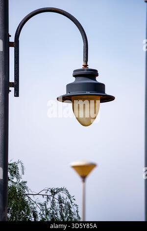 Vue verticale rapprochée d'une lanterne de rue traditionnelle de couleur foncée avec un globe blanc jaunâtre, placée contre un ciel bleu pâle, avec un second Out-o Banque D'Images