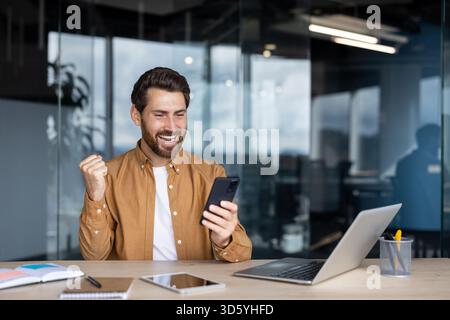 Homme souriant sentant la joie et le succès vérifier les bonnes nouvelles sur smartphone, levant le poing en triomphe tout en étant assis à un bureau dans un bureau moderne avec un ordinateur portable Banque D'Images
