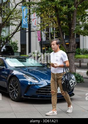Un homme décontracté au style audacieux passe devant une voiture de sport de luxe bleue garée sur un trottoir dans un quartier commerçant haut de gamme de Tokyo, au Japon Banque D'Images