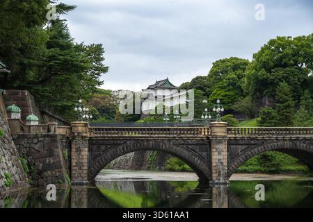 Le pont de pierre historique enjambe les douves menant au parc du Palais impérial de Tokyo, encadré par des arbres verts et une architecture japonaise traditionnelle. Banque D'Images