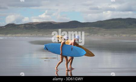 Un couple, portant tous deux des planches de surf, marche pieds nus sur le sable noir humide de Wainui Beach, en Nouvelle-Zélande Banque D'Images