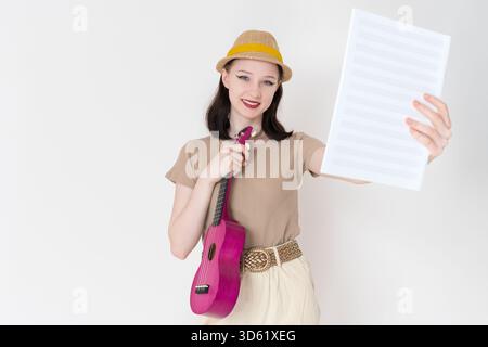 Jeune femme joyeuse joueur de ukulélé tient ukulélé et partition dans la main tendue et regarde la caméra avec le sourire. Heureuse belle musicienne Banque D'Images