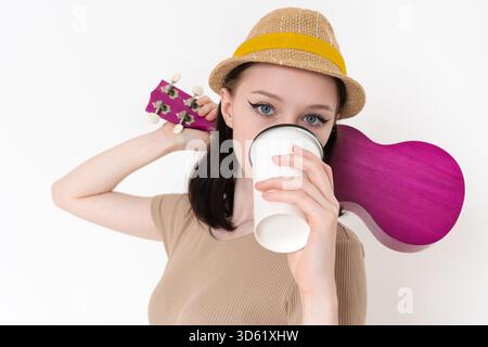 Jeune femme musicienne et joueur de ukulélé buvant du café de la tasse de papier blanc pendant sa pause café à la répétition. Une jeune femme caucasienne tient du ukulélé Banque D'Images