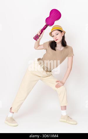 Jeune musicienne hipster et danseuse tenant ukulélé au-dessus de sa tête, se tient debout avec les jambes écartées et une main sur sa hanche. Jeune femme de Genera Banque D'Images
