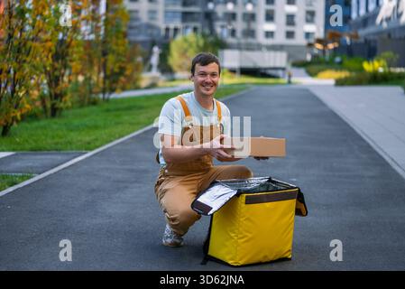 Livreur souriant en salopette manipulant soigneusement les colis en carton, les plaçant dans ou les récupérant d'un sac thermique jaune, reflétant les services de livraison urbains modernes et pratiques Banque D'Images