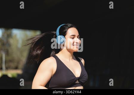 Une jeune femme forte fait du sport et court dans le parc heureux, écoutant de la musique avec des écouteurs Banque D'Images