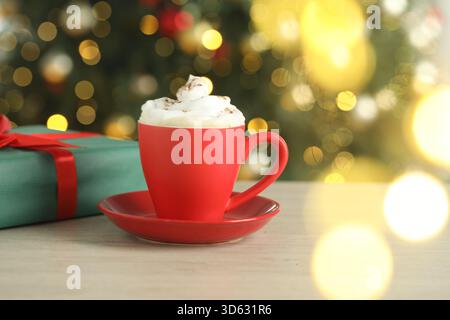 Café aromatique avec crème fouettée et cadeau de Noël sur la table en bois blanc contre les lumières floues, gros plan Banque D'Images