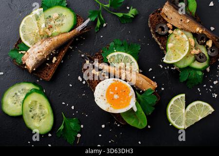 Sandwichs de poisson avec sprats, concombre, citron vert, oeufs durs, feuilles de persil et bette sur pain de seigle sur fond noir vieux béton ou pierre. Mise au point sélective. Style rustique. Vue de dessus. Copier l'espace. Banque D'Images