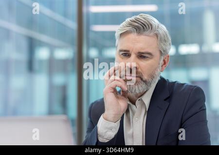 Homme d'affaires barbu mature regardant un écran d'ordinateur portable, se concentrant sur un problème sérieux, réfléchissant à un défi et trouvant une solution dans un environnement de bureau moderne Banque D'Images