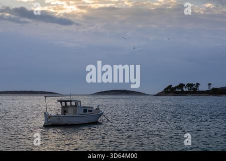Paysage côtier tranquille avec un petit bateau sur l'eau bleue calme, une île lointaine, et un village de pierre pittoresque le long du rivage sous un ciel nuageux Banque D'Images