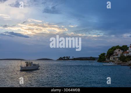 Paysage côtier tranquille avec un petit bateau sur l'eau bleue calme, une île lointaine, et un village de pierre pittoresque le long du rivage sous un ciel nuageux Banque D'Images