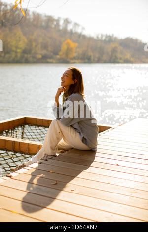 Une jeune femme se détend sur un quai en bois au bord d'un lac serein, se prélasser dans le soleil chaud de l'après-midi, entouré par la nature. Banque D'Images