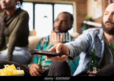 Gros plan sur l'homme caucasien à l'aide de la télécommande, ajustant le volume de la télévision pendant que les amis s'assoient sur le canapé à proximité. Blanc barbu changeant de chaîne, choisissant le divertissement pendant le repaire à la maison. Banque D'Images