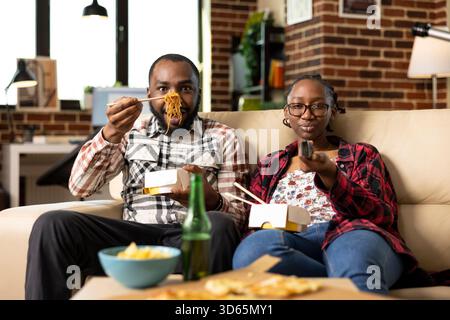Couple afro-américain se relaxant à la maison, partageant des plats à emporter sur le canapé. L'homme utilise des baguettes pour manger des nouilles que la femme feuillette à travers les chaînes de télévision, profitant d'une soirée confortable et décontractée ensemble. Banque D'Images