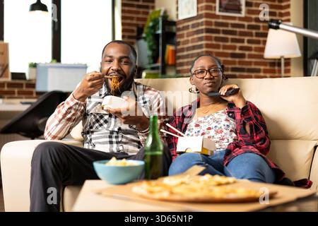Homme barbu appréciant les nouilles comme petite amie tient à distance, à la fois la liaison sur l'émission de télévision dans l'appartement de mur de briques. Jeune couple noir ayant une ambiance détendue week-end sur le canapé confortable du salon. Banque D'Images