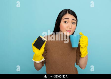 Jolie femme nettoyante portant des gants en caoutchouc jaune tient la tasse à café à la bouche, prenant la pause des tâches du matin. Femme de ménage asiatique debout avec éponge de nettoyage et tasse à la main, profitant d'un bref repos. Banque D'Images