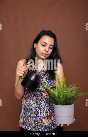 Femme indienne arrose soigneusement la petite plante verte, debout isolé sur fond brun. Idéal pour le contenu axé sur le jardinage d'intérieur, l'entretien des plantes et la conscience environnementale. Banque D'Images
