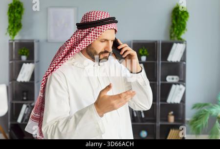 Homme d'affaires arabe portant la robe traditionnelle de dishdasha debout dans le bureau parlant par téléphone portable. Banque D'Images