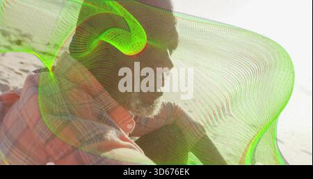 Assis homme mature regardant droit sur le rivage de sable, portant chemise à carreaux, casquette, superposition de maille néon Banque D'Images