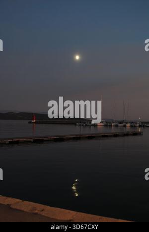 Voiliers la jetée et la tour de lumière devant les nuages orange rosé au coucher du soleil avec des collines de pleine lune et leurs reflets sur l'eau Banque D'Images