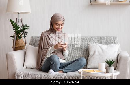 Femme arabe heureuse en hijab à l'aide d'un smartphone sur un canapé à la maison Banque D'Images
