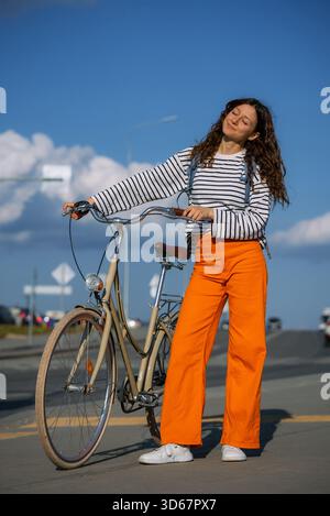 Femme debout avec un vélo de ville sur une rue urbaine ensoleillée, souriante et poussant entre les promenades - navette écologique sur une journée détendue et active embrassant un style de vie sain en plein air et la liberté Banque D'Images