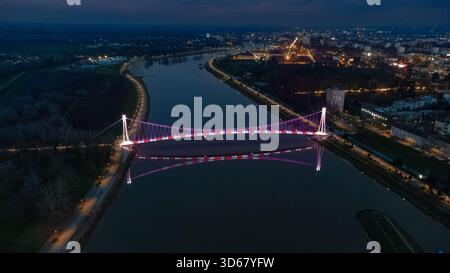 Vue aérienne du pont piétonnier illuminé enjambant la paisible rivière Drava, reflétant les lumières de la ville dans un spectacle fascinant, Osijek, Osije Banque D'Images