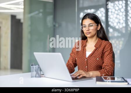 Jeune femme d'affaires millénaire portant des lunettes assis à un bureau et tapant sur un ordinateur portable, se concentrant tout en travaillant sur un projet dans un environnement de bureau d'entreprise Banque D'Images