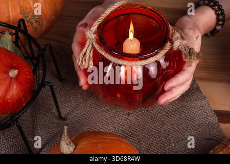 Une personne tient un bougeoir décoratif en forme de citrouille avec une bougie allumée à l'intérieur. Une citrouille orange est placée à proximité sur une table en bois Banque D'Images