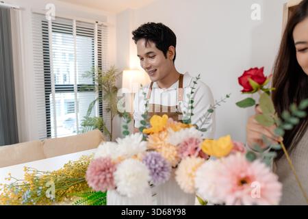 Un jeune couple arrangeant des fleurs colorées ensemble dans un cadre de maison confortable. L'homme, portant un tablier, aide la femme à créer un beau bouquet, shar Banque D'Images