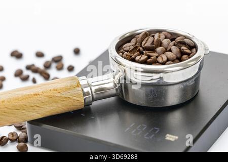 Gros plan d'un porte-filtre rempli de grains de café torréfiés reposant sur une balance à café numérique affichant un poids de 18,0 grammes, avec des grains dispersés A. Banque D'Images