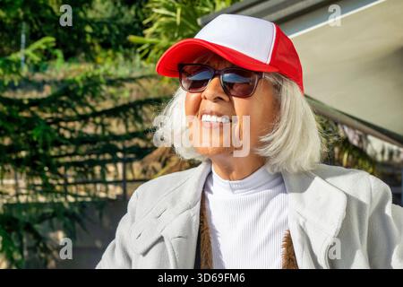 Portrait d'une femme âgée heureuse avec les cheveux blancs portant des lunettes de soleil et une casquette rouge tout en se tenant à l'extérieur par une journée ensoleillée. Concept de vieillissement actif, HAP Banque D'Images