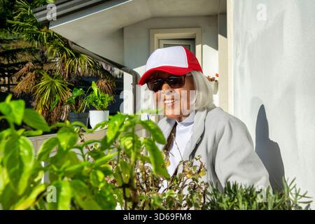 Femme âgée heureuse portant des lunettes de soleil et une casquette rouge tout en jardinant sur son balcon par une journée ensoleillée. Concept de mode de vie actif, vieillissement sain, Happine Banque D'Images