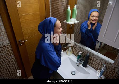Une femme arabe musulmane dans un hijab bleu sourit au miroir de la salle de bain tout en touchant son visage, avec des bouteilles et des articles d'évier à proximité, illustrant son état de vie quotidien Banque D'Images