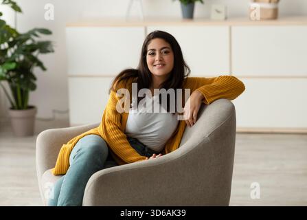 Heureuse femme arabe se détend dans un fauteuil confortable lors d'un week-end paisible matin à la maison Banque D'Images