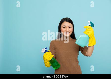 Concierge asiatique excité et souriant tenant des sprays de nettoyage dans des gants en caoutchouc jaunes, prêt à commencer les tâches quotidiennes. Femme au foyer optimiste avec des désinfectants, montrant une attitude positive envers l'entretien ménager. Banque D'Images