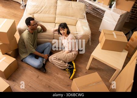 Couple diversifié est assis sur le sol après le jour de déménagement, se détendre et parler ensemble dans un appartement confortable, entouré de boîtes en carton. Petit ami et petite amie se lient tout en profitant de la nouvelle maison. Banque D'Images