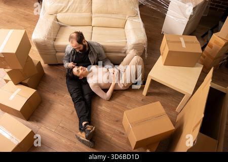 Le jeune couple aime le collage sur le sol du salon pendant le jour du déménagement. Belle femme se repose sur les genoux du partenaire, entourée de meubles et de boîtes en carton, embrassant la relocalisation et la propriété d'une nouvelle maison. Banque D'Images