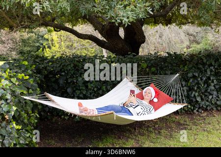 Femme s'inclinant sur un hamac sous l'arbre dans le jardin portant chapeau de Père Noël et chaussettes rouges tenant smartphone Banque D'Images