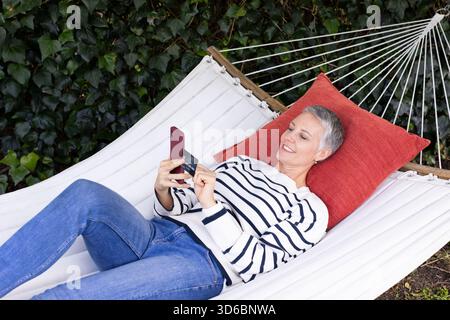 Femme senior s'inclinant sur hamac de toile dans la cour arrière tenant le téléphone et la carte de crédit avec coussin rouge Banque D'Images