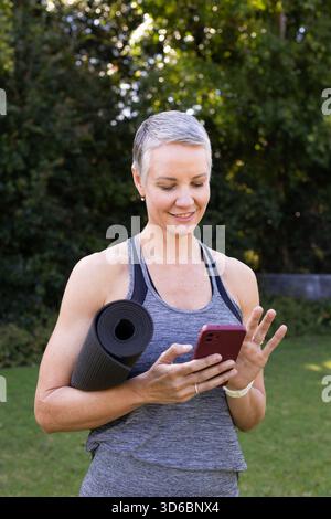 Femme senior portant un débardeur, debout dans la cour tenant un tapis de yoga noir, à l'aide d'un smartphone rose Banque D'Images