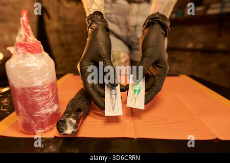 Tatoué jeune homme adulte caucasien portant des gants noirs tenant deux aiguilles de tatouage emballées sur la surface orange, se préparant pour la procédure de tatouage dans le cadre du studio Banque D'Images