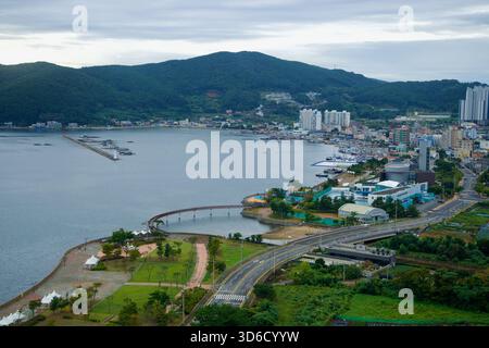 Le port de Jisepo dans la ville de Geoje s'étend en contrebas, avec le parc côtier et la promenade courbe au premier plan, des bateaux de pêche amarrés le long du brise-lames Banque D'Images