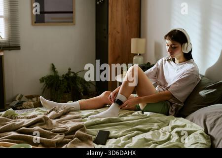 Jeune femme biraciale assise à l'intérieur portant un moniteur de cheville et écoutant de la musique Banque D'Images