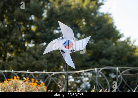 Une roue à épingles blanche décorée de drapeaux sud-coréens tourne dans la brise près d'une clôture en fil de fer barbelé, sur fond d'arbres et de lumière du soleil Banque D'Images