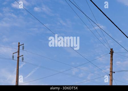 Deux types différents de poteaux électriques, l'un moderne et l'autre plus ancien, se dressent contre un ciel bleu avec des nuages légers, leurs lignes électriques croisées. Banque D'Images